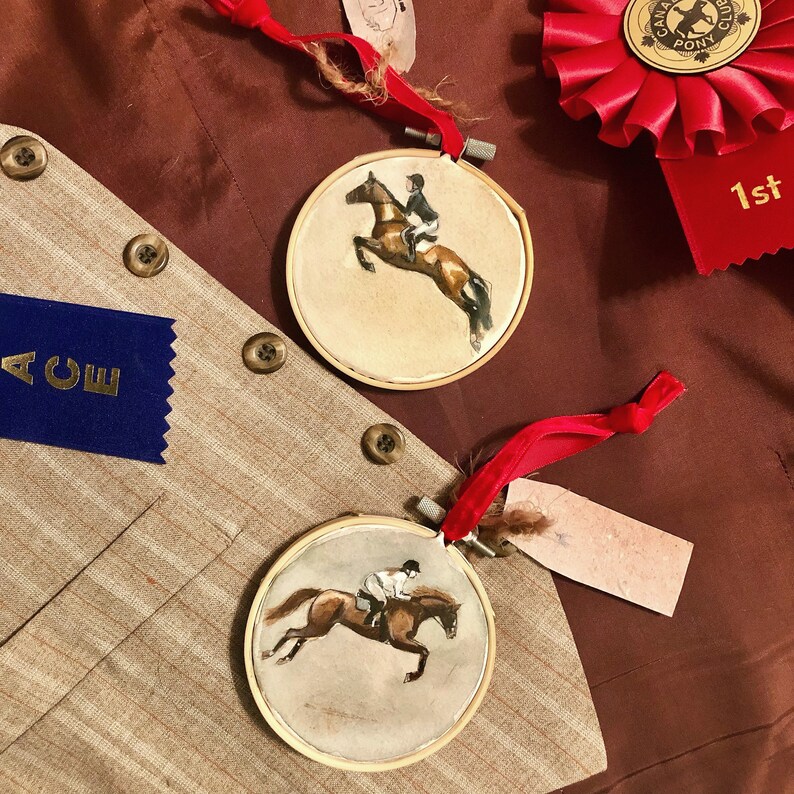 K&ouml;nnte beinhalten: Zwei runde, gerahmte Aquarellmalereien von Springreiten, jeweils mit einem roten Band. Die Gem&auml;lde zeigen einen Reiter auf einem braunen Pferd, das &uuml;ber ein Hindernis springt. Ein blaues Band mit dem Wort "ACE" ist sichtbar.