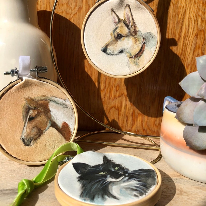 K&ouml;nnte beinhalten: Aquarellportr&auml;ts eines Hundes, Pferdes und einer Katze in Holz-Stickrahmen. Der Hund hat braunes und wei&szlig;es Fell, das Pferd ist braun und wei&szlig;, und die Katze ist schwarz und wei&szlig;. Ein gr&uuml;nes Band ist befestigt.