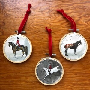 K&ouml;nnte beinhalten: Drei runde Ornamente mit aquarellierten Gem&auml;lden zum Thema Reiten. Jedes Ornament zeigt ein Pferd und einen Reiter oder ein Pferd mit einer Decke. Die Ornamente sind in Holzreifen gefasst und haben rote Samtbandschlaufen.