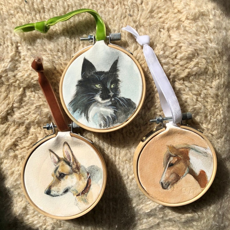 K&ouml;nnte beinhalten: Drei runde Stickrahmen mit Aquarellportr&auml;ts von Tieren. Ein schwarz-wei&szlig;er Katze, ein Hund und ein Pferd sind abgebildet. Jeder Rahmen hat ein Band zum Aufh&auml;ngen.