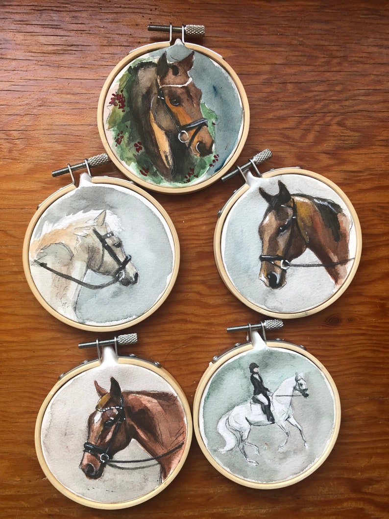 K&ouml;nnte beinhalten: F&uuml;nf Aquarellmalereien von Pferden in Stickrahmen. Die Pferde sind in verschiedenen Farben dargestellt, darunter Braun, Wei&szlig; und Grau. Jedes Pferd tr&auml;gt ein Zaumzeug. Ein Gem&auml;lde zeigt einen Reiter auf einem wei&szlig;en Pferd. Die Reifen sind aus Holz.