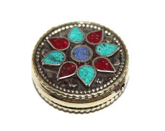 May include: A round, silver-colored trinket box with a floral design. The flower is made of red, turquoise, and blue stones. The box has a gold-colored rim and a detailed edge.