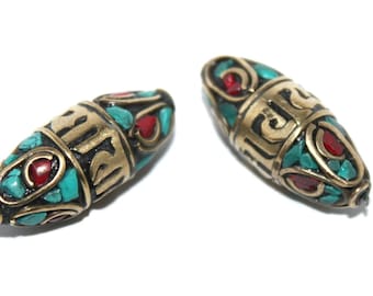 May include: Two oval-shaped beads with intricate designs. The beads are made of brass and feature turquoise and red accents. The beads are decorated with a repeating pattern of Sanskrit characters.