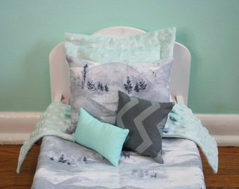 May include: A miniature doll bed with a white headboard, featuring a gray and white landscape-printed pillow and blanket set. Additional pillows include a teal polka-dot pillow and a gray chevron-patterned pillow. The bedding has a soft, quilted texture.
