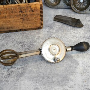 May include: An antique hand-crank egg beater with a black wooden handle and a silver-colored whisk. The beater is on a gray surface, along with other vintage kitchen tools, including a grater and a muffin tin. A wooden crate is in the background.