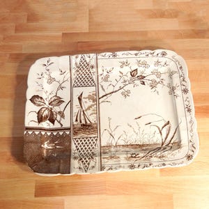 May include: Rectangular serving tray with a scalloped edge. The tray features a brown and white landscape design with floral and nautical elements. The tray is made of ceramic or porcelain.