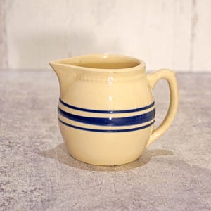 May include: Cream-colored ceramic pitcher with a spout and handle, featuring two horizontal blue stripes around the body. The pitcher has a classic, vintage design, suitable for serving beverages or as a decorative item.