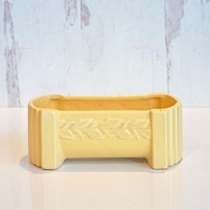 May include: A rectangular, pale yellow ceramic planter with a decorative leaf pattern. The planter has a low profile with a slightly curved top edge and vertical ribbed sides. The planter is approximately 8 inches long.