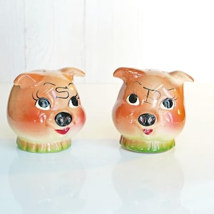 May include: A pair of vintage pig-shaped salt and pepper shakers. The ceramic shakers are tan with pink cheeks, black eyes, and red lips. The letter "S" is on one pig and "P" on the other. The base of each shaker is green.