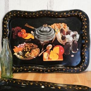 May include: A black metal serving tray with a gold border and a floral design. The tray features a colorful illustration of a variety of food and drinks, including a chafing dish, cheese, fruit, and bottles of Coca-Cola.