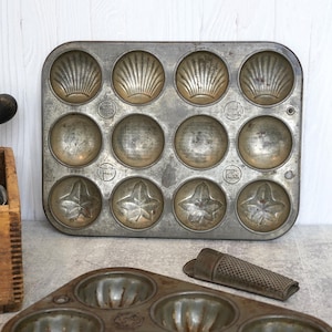 May include: Vintage metal muffin tin with seashell, circle, and leaf-shaped molds. The tin has a worn, aged appearance, suggesting it's been well-used. A wooden box with kitchen tools and a grater are also visible.