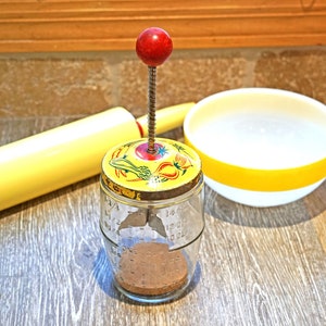 May include: A vintage yellow and red manual food mill with a glass jar and a red knob on top. The mill is sitting on a wooden surface next to a yellow rolling pin and a white and yellow bowl.