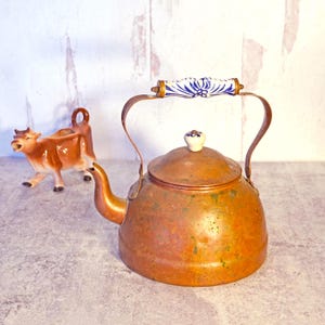 May include: A copper tea kettle with a decorative handle and lid finial. The handle is white with blue floral designs. A ceramic cow creamer is in the background. The kettle has a spout and a curved handle.