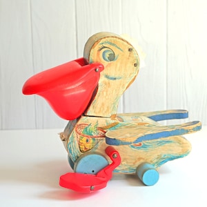 May include: Vintage wooden pelican pull toy with a red beak and feet, blue accents, and painted details. The toy has wheels and is designed to be pulled along a surface. A classic children's toy.