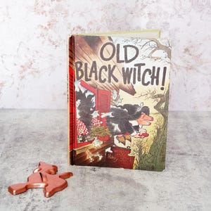 May include: A hardcover book titled Old Black Witch! by Wende and Harry Devlin, featuring an illustration of a witch on a broomstick flying past a house, accompanied by a copper-colored metal witch-shaped cookie cutter on the surface.