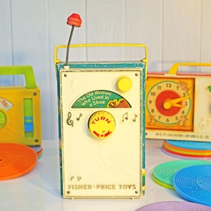 May include: Three vintage Fisher-Price record players with colorful plastic cases and spinning records. The middle record player features a yellow button and a red knob with the text "The Old Woman Who Lived in a Shoe" and "TURN".