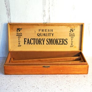 May include: Open wooden box with the text "FACTORY SMOKERS" and "FRESH QUALITY" printed on the inside lid. The box is a light brown color and has a small metal clasp. The text also includes the price of 5 cents.