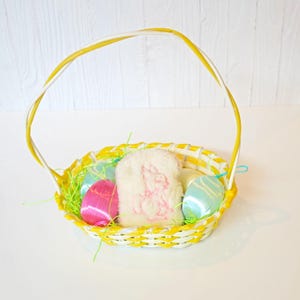 May include: A yellow and white woven Easter basket filled with colorful eggs and decorative grass. A white, fluffy bunny-shaped item is nestled among the eggs. The basket has a yellow handle and sits on a white surface.
