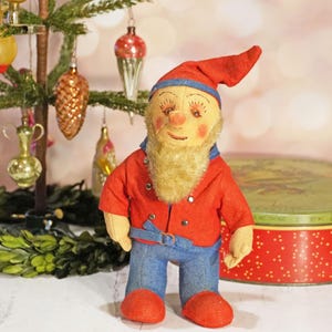 May include: A vintage, handmade, red and blue felt gnome doll with a white beard and a red hat. The gnome is standing in front of a Christmas tree and a red and green tin.