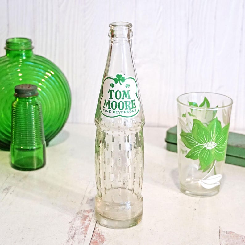 May include: A clear glass Tom Moore Fine Beverages bottle with a green shamrock logo. The bottle is next to a green glass with a floral design and other green glass objects.