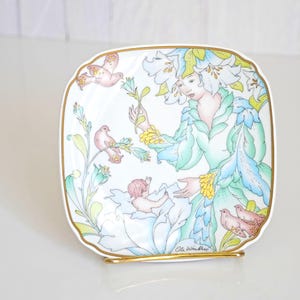 May include: A square decorative plate with a gold rim and a gold stand. The plate features a painted design of a woman surrounded by flowers, birds, and cherubs in pastel colors. The artist's signature is visible on the plate.