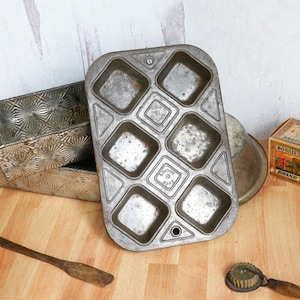 May include: Vintage metal muffin tin with eight square molds, showing signs of wear. Beside it are other vintage baking items, including a loaf pan and a small box. The items are on a wooden surface.