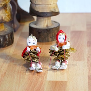 May include: Two vintage Christmas ornaments depicting children skiing. The ornaments are made of porcelain and pine cones, with red and white accents.