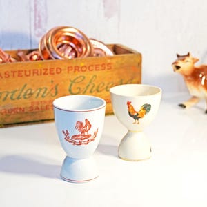 May include: Two white ceramic egg cups with rooster designs. One cup has a red rooster illustration, the other a colorful rooster. A wooden box with text and copper rings are in the background.