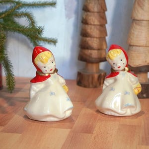 May include: Two ceramic figurines of girls wearing red hooded cloaks and white dresses. The girls are holding baskets in their hands. The figurines are standing on a wooden surface.