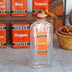 May include: A clear glass bottle with a red lid and a label that reads "TONE'S Imitation Coconut Flavor". The bottle is sitting on a table with other spice jars in the background.