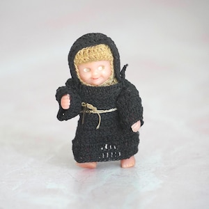 May include: A small doll dressed in a black crocheted monk's habit with a hood. The doll has blonde hair and a serene expression. A beige cord is tied around the waist. The doll is standing on a light-colored surface.