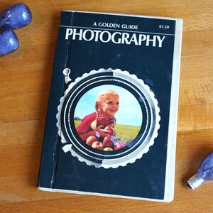 May include: A black and white book titled "A Golden Guide Photography" with a photo of a young girl holding apples on the cover. The book is priced at $1.50.