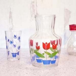 May include: Clear glass pitcher and tumbler set decorated with red tulip designs. The pitcher has a wide base and a narrow neck. The tumbler has a straight shape. The tulips are in blue pots with green leaves. A red-topped container is also visible.