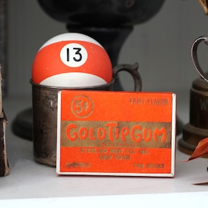 May include: A vintage box of Gold Top Gum, fruit flavored, with a red and white billiard ball with the number 13 on it sitting on top of the box.