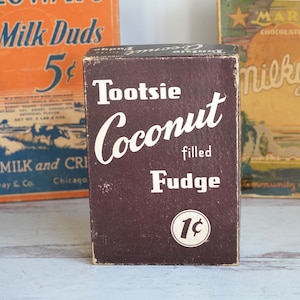 May include: A vintage Tootsie Coconut Fudge candy box with a brown background and white text. The box reads "Tootsie Coconut filled Fudge 1c".