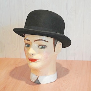 May include: A classic black bowler hat with a rounded crown and a short, curled brim. The hat is constructed from a dark, felt-like material and features a matching black ribbon band encircling the base of the crown, resting above the brim.