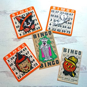 May include: Five Halloween-themed bingo cards with orange and white backgrounds. Each card features a different spooky character: a black cat, a ghost, a vampire, a witch, and a jack-o'-lantern. The cards are printed with the word "Bingo" and numbers.