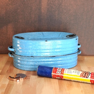 May include: Two stacked, light blue enamelware oval baking dishes with black handles. A blue and red can-shaped item with text is next to a copper-colored bottle opener. The items are on a wooden surface.