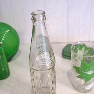 May include: Clear glass bottle with text, a green glass tumbler with a floral design, and other green glass containers. The bottle has a textured base and a narrow neck. The tumbler features a green and white floral pattern.