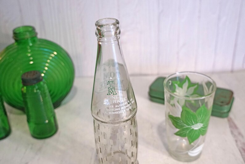 May include: Clear glass bottle with text, a green glass tumbler with a floral design, and other green glass containers. The bottle has a textured base and a narrow neck. The tumbler features a green and white floral pattern.