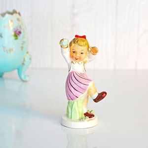May include: A vintage porcelain figurine of a young girl in a white top, pink striped skirt, and red bow. The girl is posed mid-dance on a green base. A light blue egg-shaped vase with gold accents and floral details is in the background.