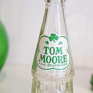 May include: Clear glass bottle with a green and white label. The label features the text "TOM MOORE FINE BEVERAGES" and a shamrock. The bottle is marked "10 FLUID OZ".