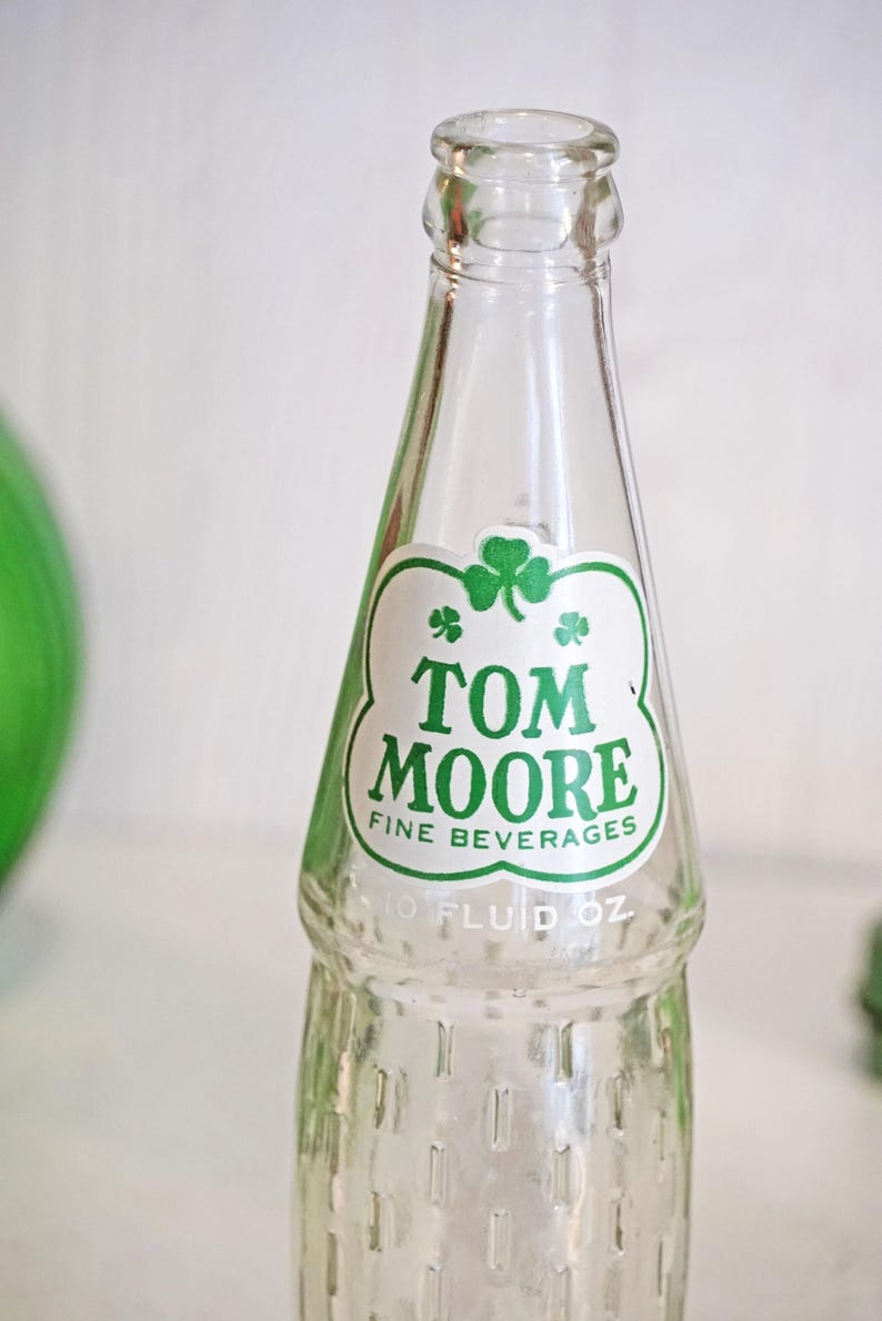 May include: Clear glass bottle with a green and white label. The label features the text "TOM MOORE FINE BEVERAGES" and a shamrock. The bottle is marked "10 FLUID OZ".