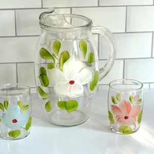 May include: A set of three glass pitcher and glasses with hand-painted floral designs. The pitcher has a white flower with green leaves, one glass has a light blue flower, and the other glass has a pink flower.