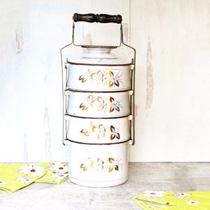 May include: Vintage white enamel tiered food carrier with a metal frame and wooden handle. Each tier is decorated with gold floral designs. The carrier is stacked on top of each other. The carrier is a vintage kitchenware item.