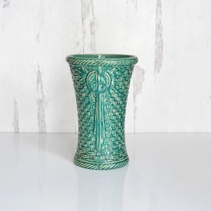 May include: A teal ceramic vase with a basket weave design. The vase has a flared top and a detailed pattern, including a central vertical element and a circular emblem. The vase is approximately 8 inches tall.