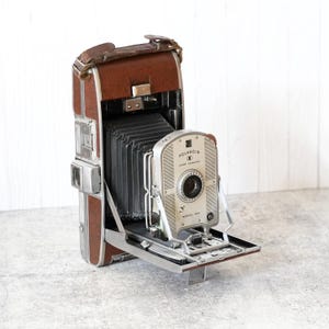 May include: A vintage Polaroid Land Camera Model 95A featuring a brown leather-covered body, a black accordion-style bellows, and a silver metal front panel with the Polaroid logo and model name clearly printed on the face.