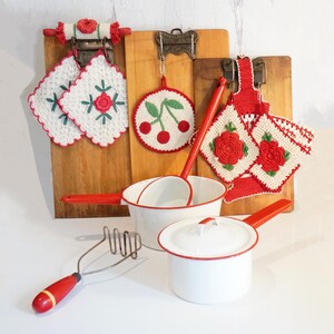May include: A collection of kitchenware including two white enamel pots with red rims and handles, a metal potato masher with a red handle, and three wooden boards displaying crocheted potholders featuring red rose and cherry motifs.