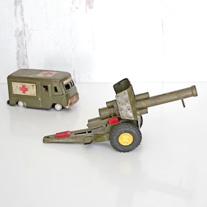 May include: Two vintage toy vehicles: an olive green ambulance with a red cross emblem and a military-style cannon. The cannon has a long barrel, yellow wheels, and red accents. Both toys are made of metal.
