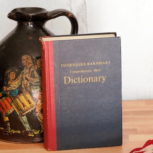 May include: A vintage black ceramic jug with a handle, featuring a painted scene of people playing drums. Beside it is a hardcover dictionary with a navy blue cover and the title "Dictionary" in gold lettering.